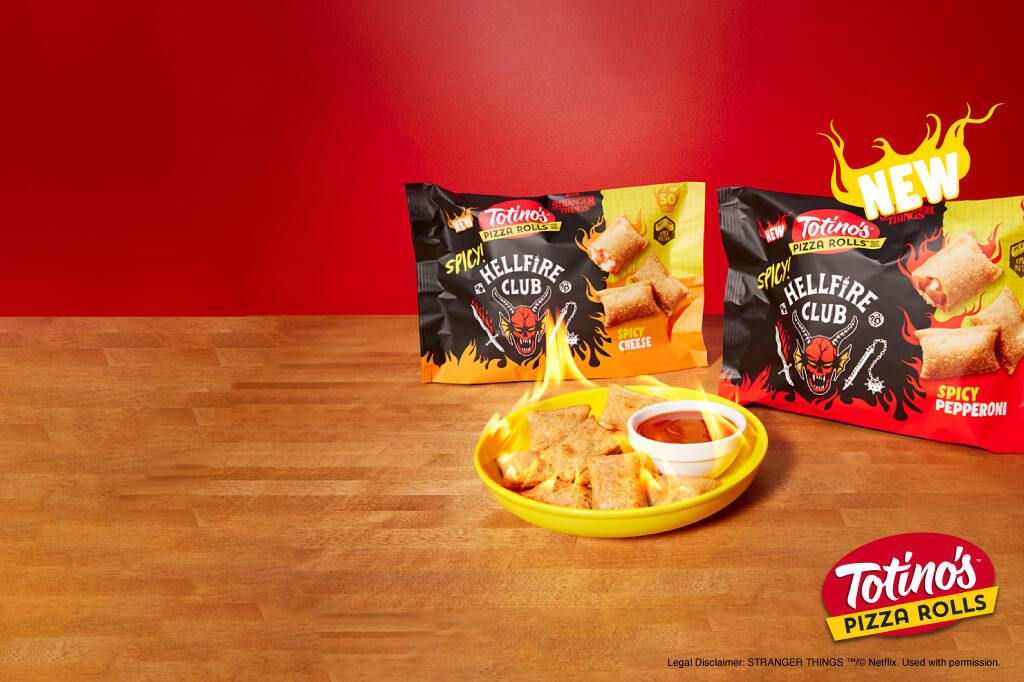 Two front facing pack shots of Totino's Pizza Rolls Spicy Hellfire Club Cheese and Spicy Hellfire Pepperoni on a wood table with a yellow plate of pizza rolls & dipping sauce in front with flames coming out the top of the plate.