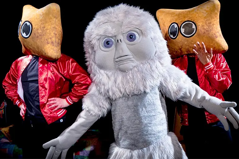Two people in red satin jackets wearing large, golden, snack-shaped mascot heads stand on either side of a fluffy white mascot character against a dark background, with a blurred rectangle centered over the middle figure.